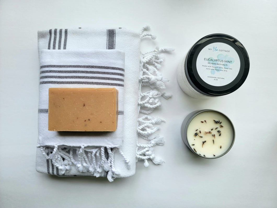 White set of Turkish bath and hand towels with tassels, a bar of soap, a eucalyptus mint bath salts, and a lavender sage candle on a white background.