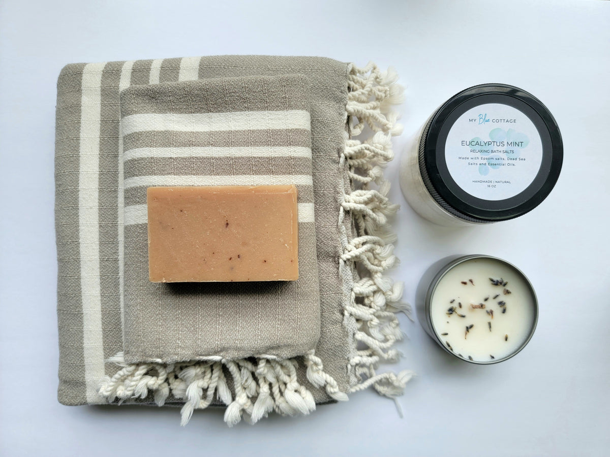 Taupe set of Turkish bath and hand towels with tassels, a bar of soap, a eucalyptus mint bath salts, and a lavender sage candle on a white background.